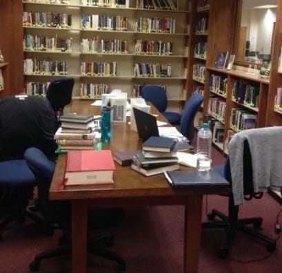 Blurry image of a large wooden table strewn with piles of books, water bottles, laptops, and pamphlets. Sweaters drape two of the chairs around it. The rest of the room is lined with bookshelves. The carpet is red.