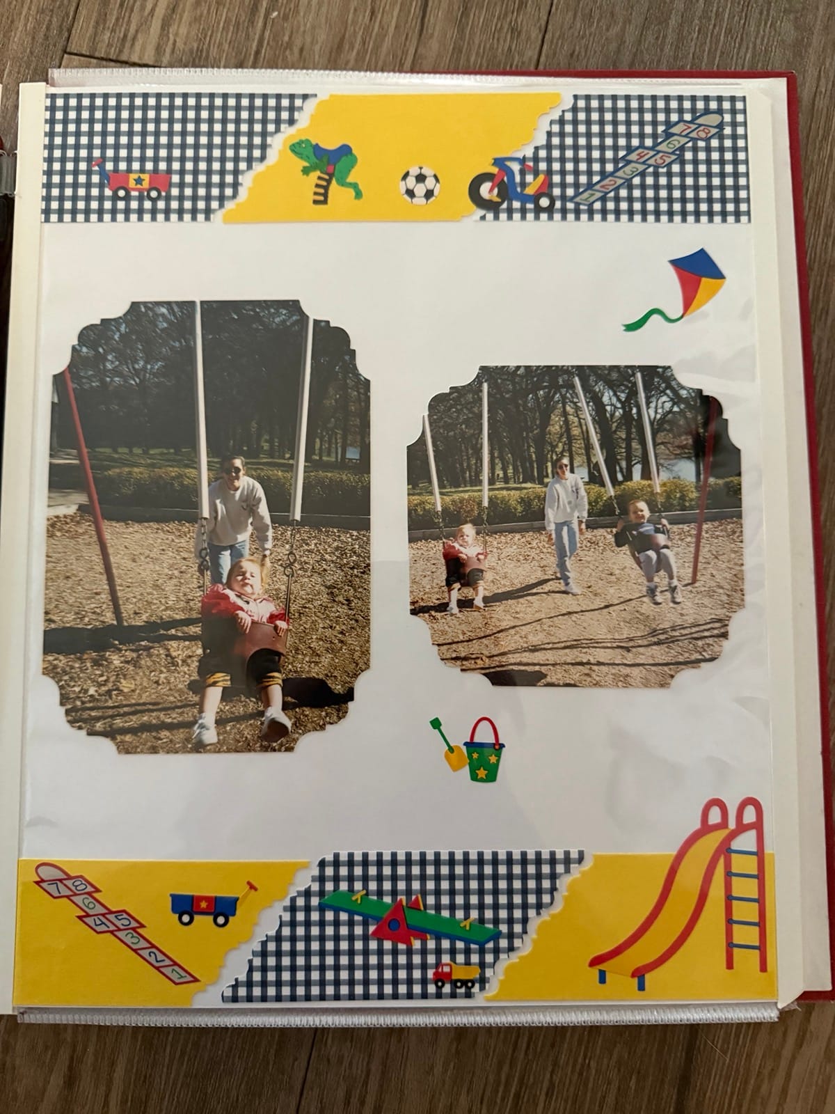 Photograph of a page of scrapbook, which features park imagery and two photos of young children swinging. The page includes stickers and a boarder of black plaid and yellow, complete with a slide, hopscotch, wagons, and other childhood accoutrements.