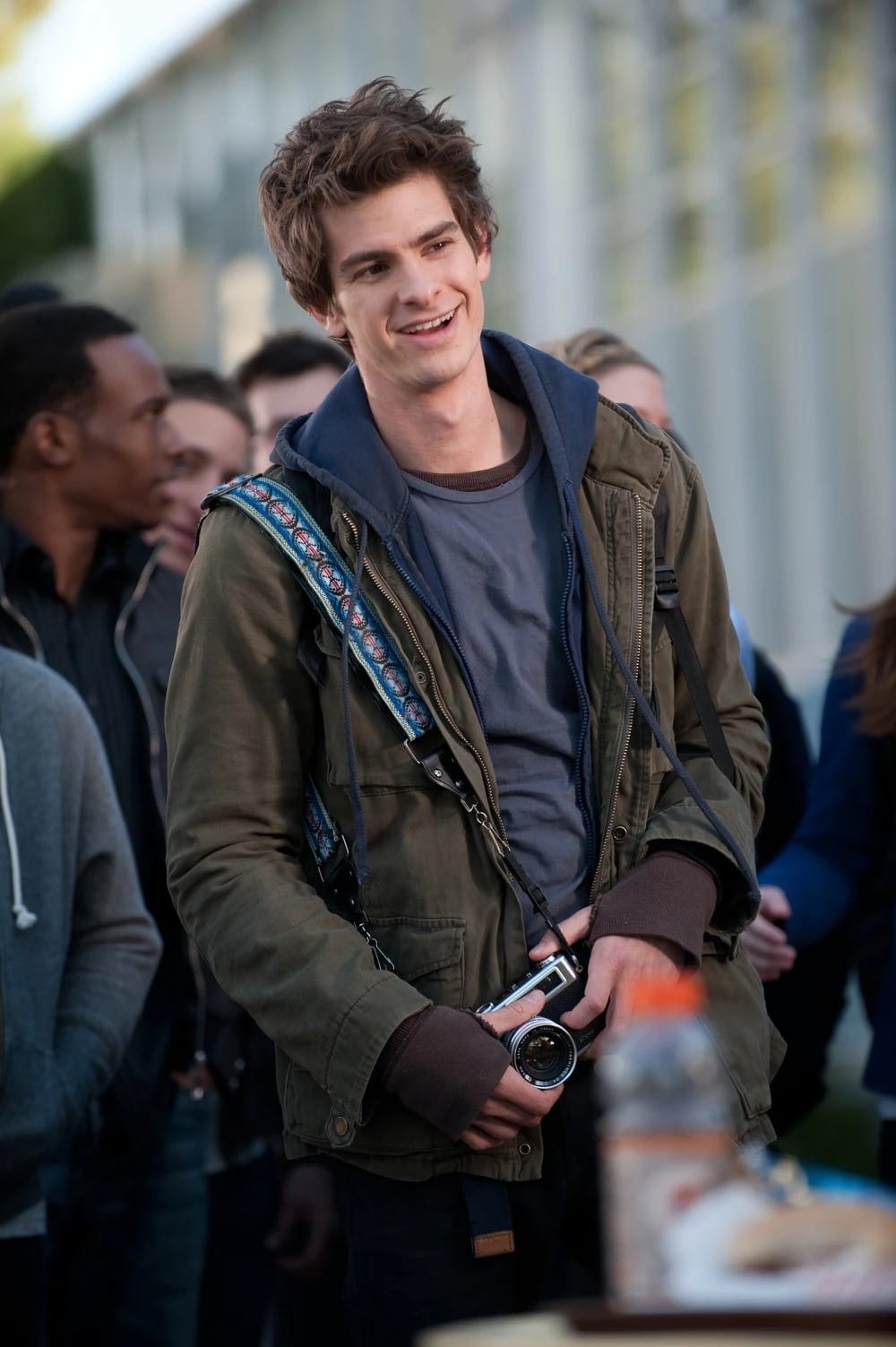 Photo of Garfield smiling as Peter Parker. He wears a brown jacket over a blue sweatshirt, blue tshirt, and maroon long-sleeve shirt with thumb holes. He holds a camera an dis surrounded by other people in the background.