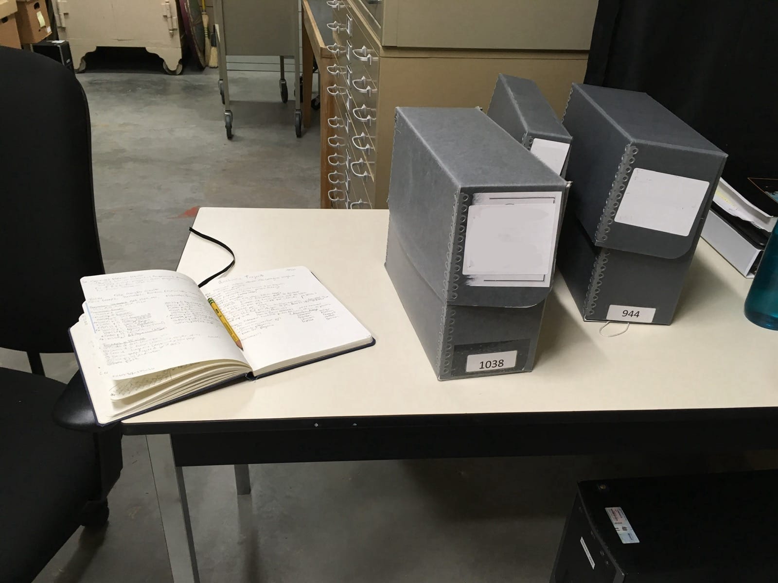 On the white-topped table is a notebook, laying open with a pencil along the crease, and three grey Hollinger boxes. A metal cabinet for oversized items stands behind the table.