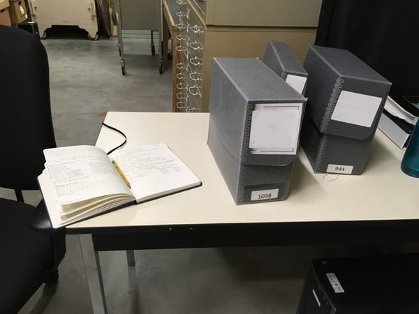 On the white-topped table is a notebook, laying open with a pencil along the crease, and three grey Hollinger boxes. A metal cabinet for oversized items stands behind the table.